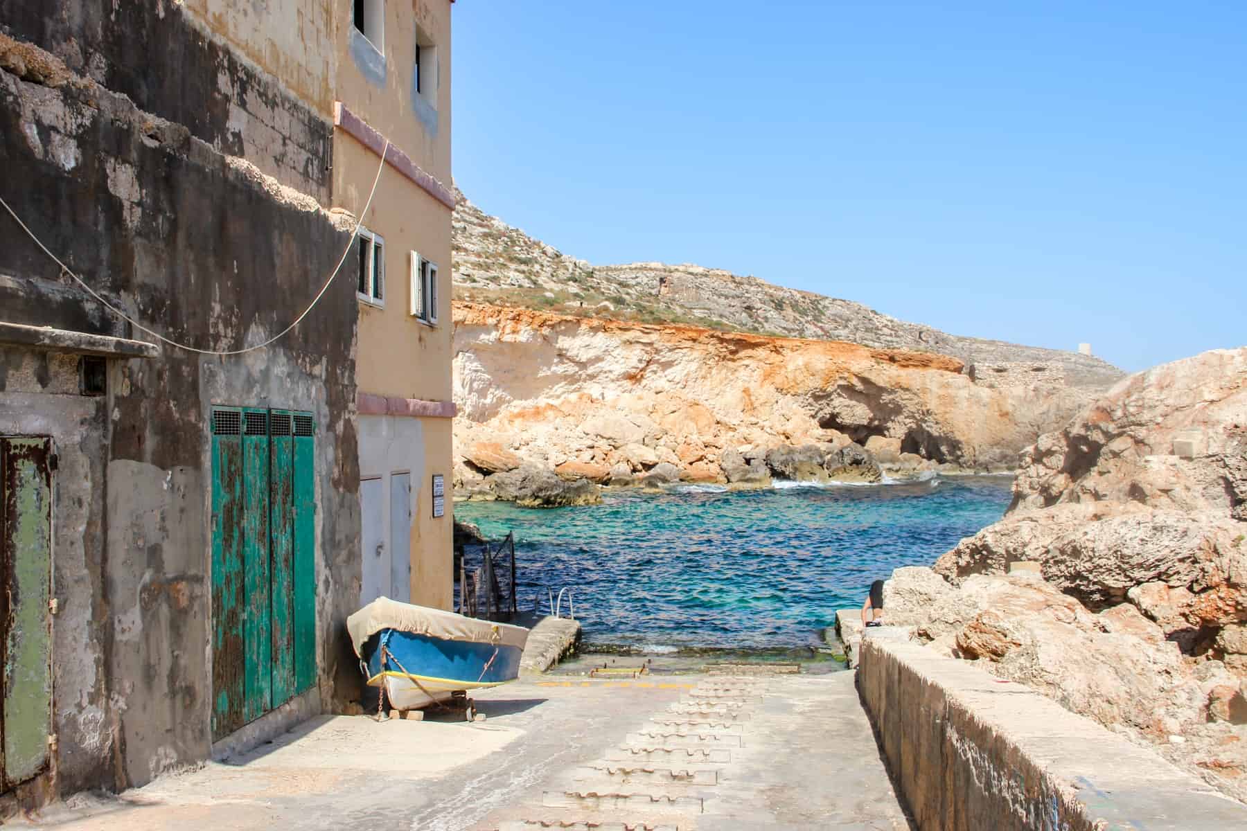 A covered boat stands next to a peach coloured building, on a slope that leads down to the blue and green sea water bay backed by orange rocks. 